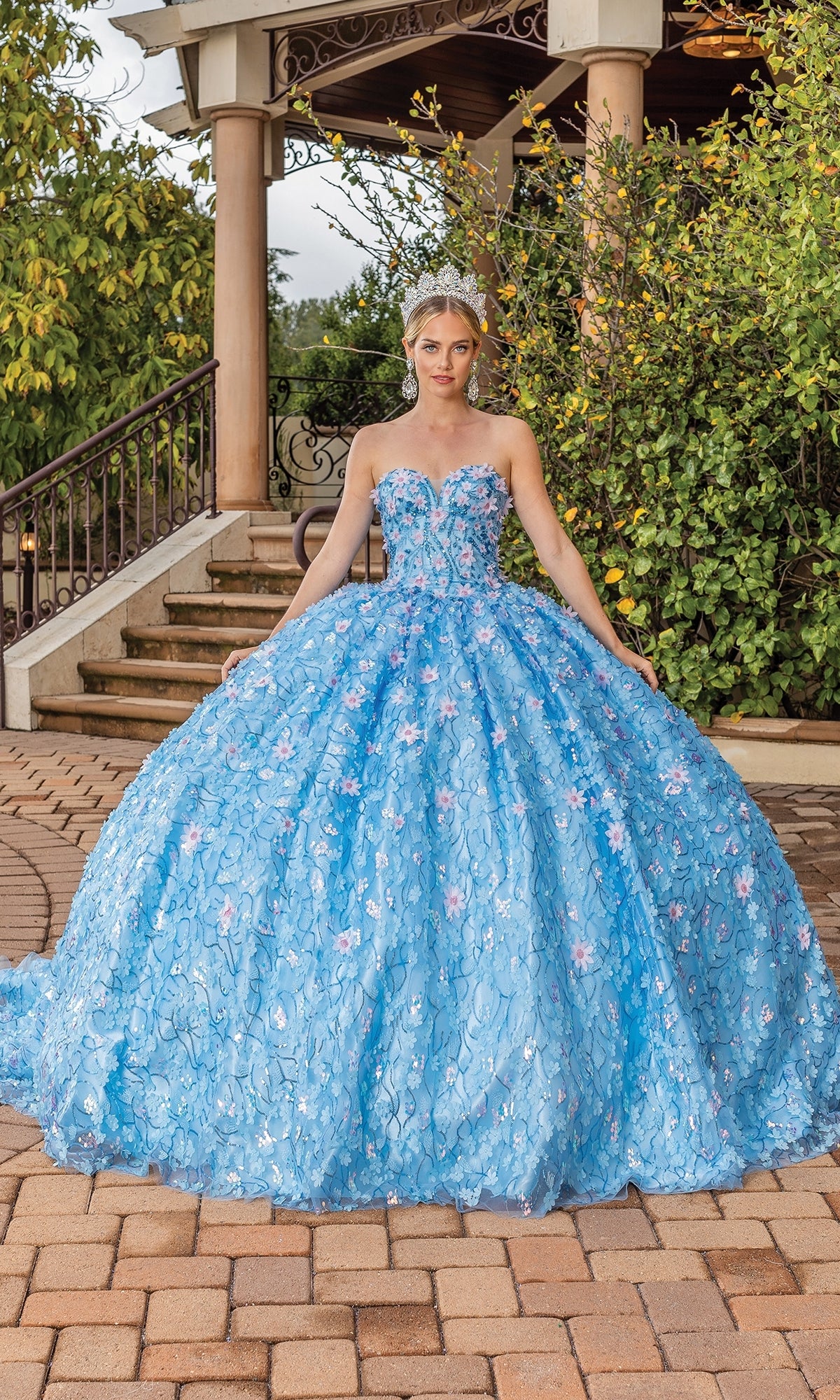 A woman stands outdoors in front of stairs, wearing the Quinceanera Dress 1835 By Dancing Queen, a blue strapless sweetheart ball gown with floral details, removable off-the-shoulder sleeves, and a sparkling tiara.